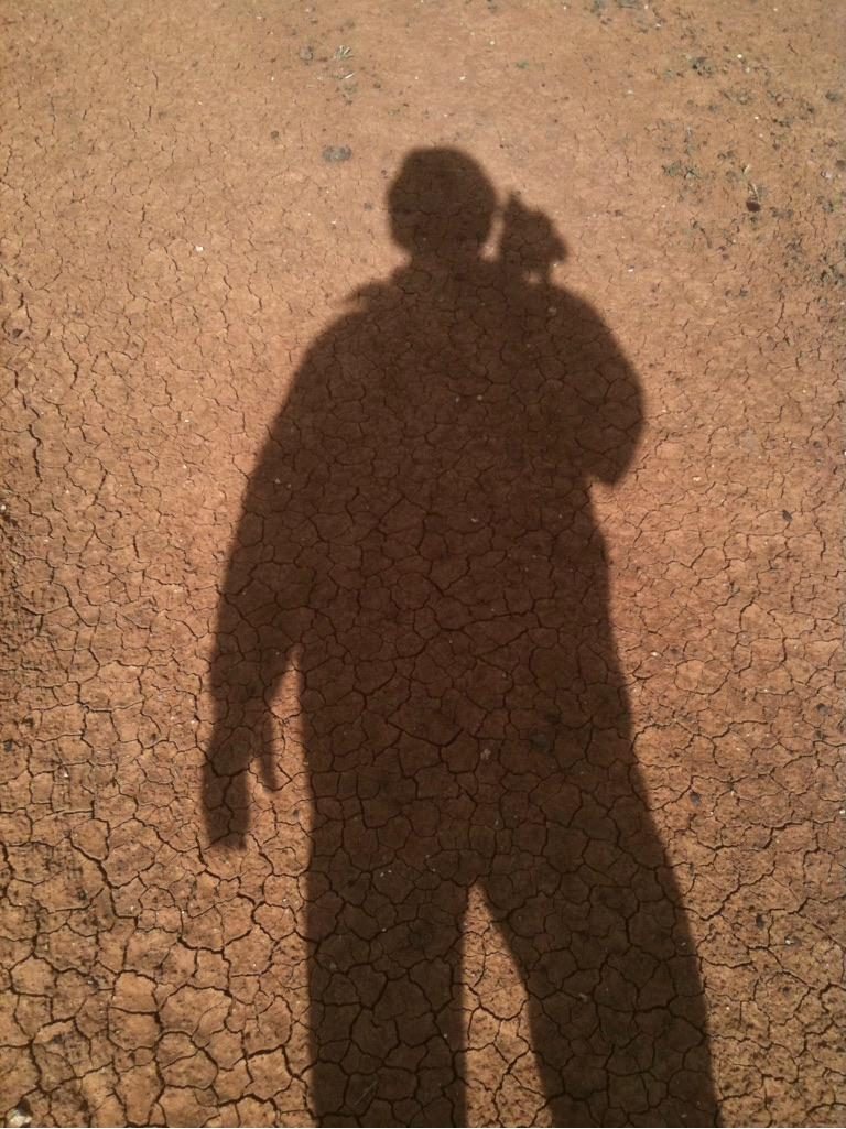The photographer's own shadow cast across dry cracked earth, with the silhouette of a small bird perched on his shoulder. Taken on a farm at Macorna, Victoria.