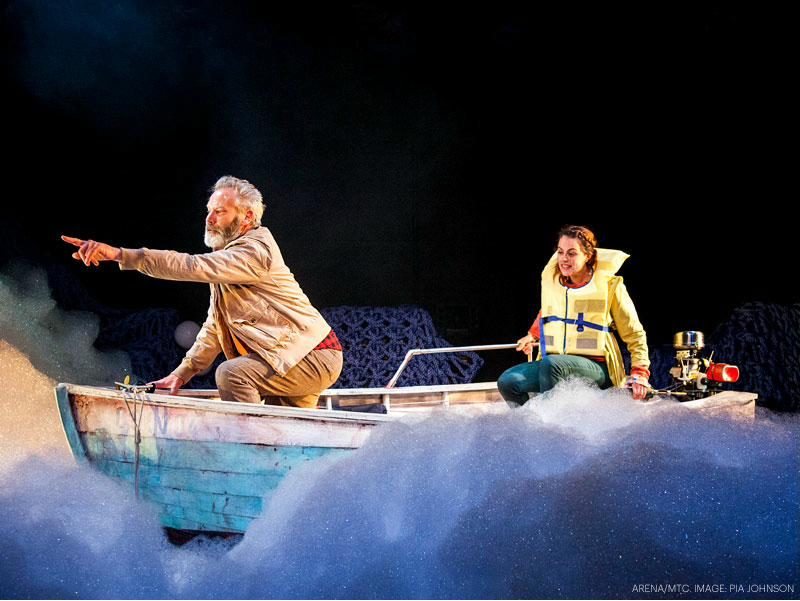 Two performers in a small wooden boat surrounded by low fog, one standing and pointing out past the audience, from the Melbourne Theatre Company and Arena Theatre Company production of Marlin.