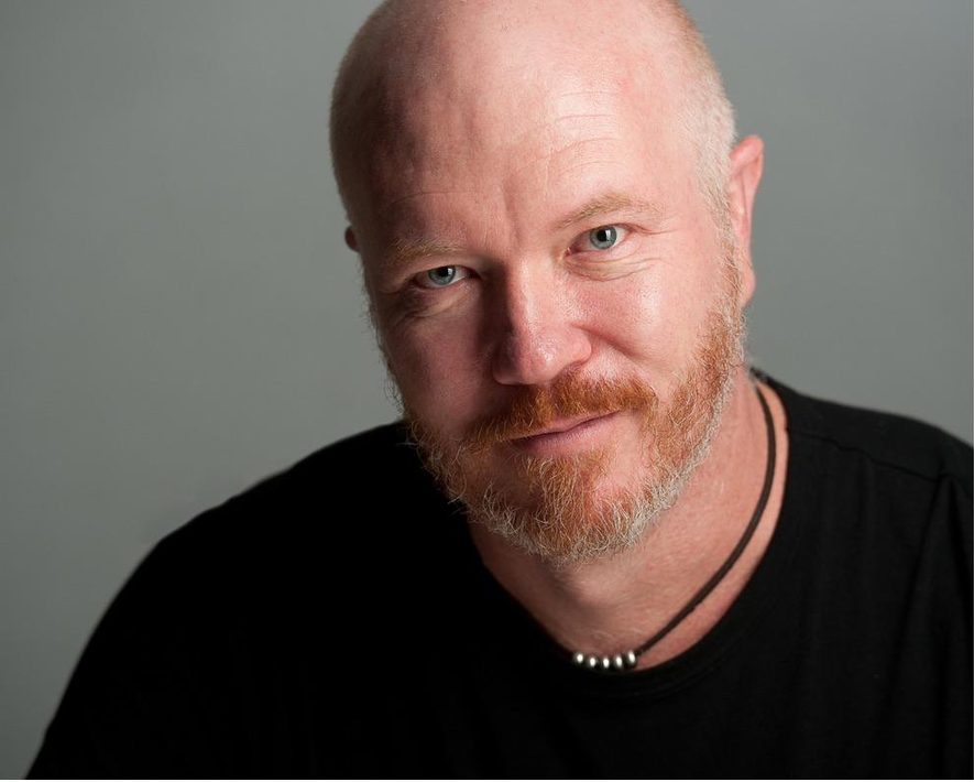 Studio portrait of Damien Millar against a grey background, close-cropped hair and a red beard, wearing a black shirt.